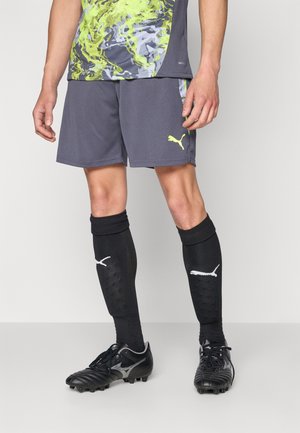Mann trägt schwarze Fußballschuhe, schwarze knielange Puma-Socken, graue Puma-Shorts und ein grün-grau gemustertes Sportshirt.