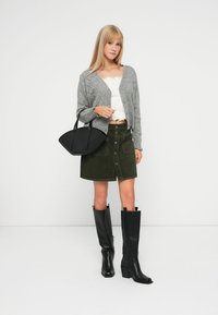 Jovem mulher vestindo um cardigan cinza, blusa branca, saia verde com botões na frente, botas pretas até ao joelho, segurando uma mala preta.