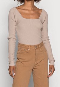 Maglia a maniche lunghe a coste di un beige chiaro con scollatura quadrata, abbinata a pantaloni marroni a vita alta con tasche e chiusura a bottone.