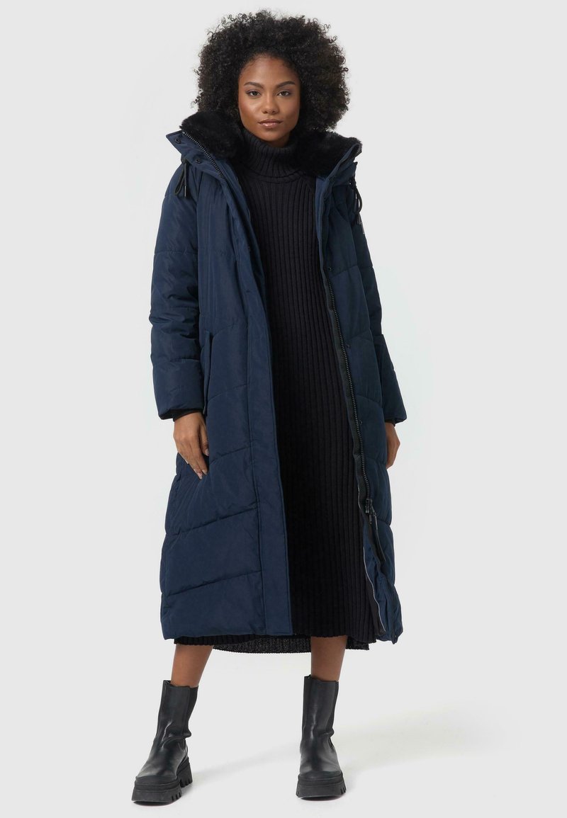 Long navy puffer coat with a fur-lined hood, featuring quilted texture, zip closure, and side pockets, worn over a black ribbed dress.