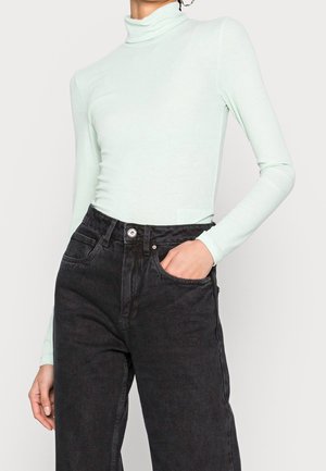 Femme portant un col roulé côtelé vert clair et un jean noir taille haute, avec une main dans la poche avant.
