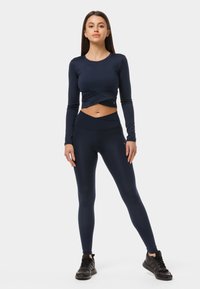 Tumedast komplitud lühikeste varrukatega crop top, millel on keerdunud esikülg, koos kõrge vöökohaga tumedate retuusidega. Mudel kannab musti sportjalatseid.