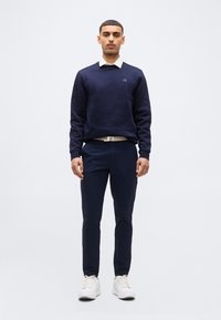 Homme debout face à l'objectif, portant un pull bleu marine par-dessus une chemise blanche à col, un pantalon bleu marine, une ceinture blanche et des baskets blanches, sur un fond uni.