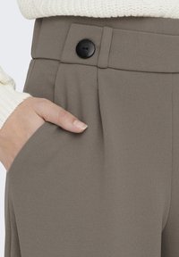 Bruine geplooide broek met een gestructureerde afwerking. Voorzien van een zwarte knoop en een zijzak, getoond met een hand die in de zak rust.