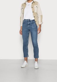 Chaqueta de botón en color beige claro con dos bolsillos en el pecho, camiseta blanca con texto, jeans cortos azules y zapatillas blancas. Denim texturizado, ajuste casual.