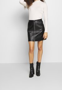 Jupe courte en cuir noir avec une fermeture éclair frontale, associée à un haut léger à manches longues côtelées et des bottines noires. Texture lisse, coupe ajustée.