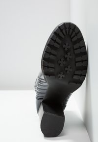 Semelle de botte noire avec des rainures profondes et des motifs en forme de fleurs, talon carré, reposant contre un fond blanc d'angle.
