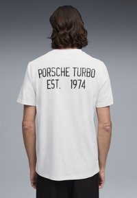 Maglietta bianca di cotone con testo nero sul retro: "PORSCHE TURBO EST. 1974." Maniche corte e una vestibilità comoda con orlo arrotondato.