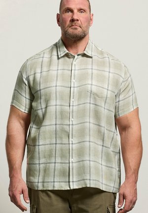 Mann trägt ein kurzärmeliges, hellgrünes kariertes Hemd mit Knöpfen und olivfarbene Cargohosen, steht vor einfachem Hintergrund.