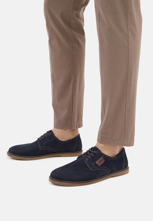 Zapatos de ante azul marino con detalles perforados, diseño con cordones y suela de goma marrón clara. Combinados con pantalones beige.