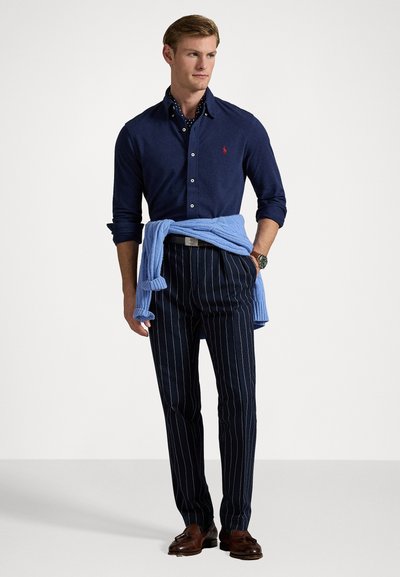 Mornarska srajca z gumbi, svetlo modro pulover obdan okoli pasu, hlače z drobnimi črtami, rjave čevlje in ure na zapestju. Priložnostna, a sofisticirana kombinacija.