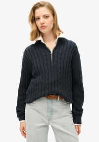 Granatowy sweter z dzianiny o wzorze warkocza, z półzamkniętym kołnierzem, ściągaczami na rękawach i luźnym krojem, połączony z jasnoniebieskimi jeansami oraz plecionym paskiem.