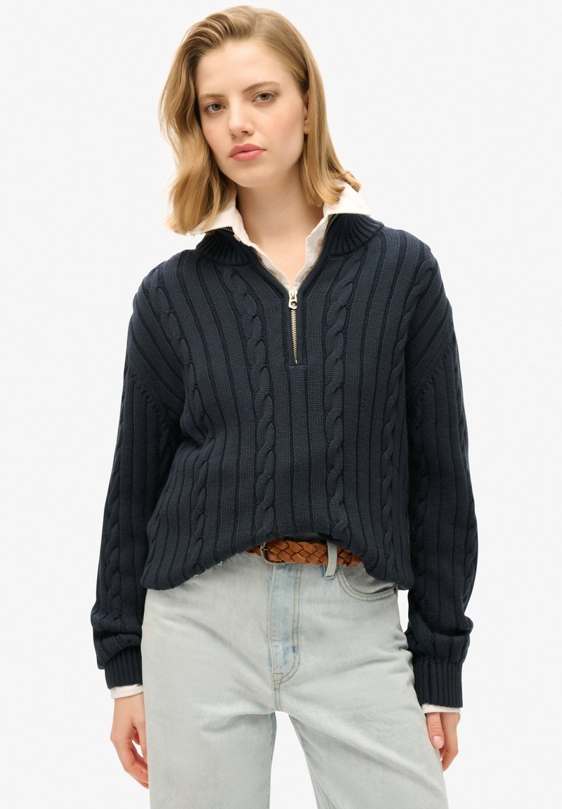 Granatowy sweter z dzianiny o wzorze warkocza, z półzamkniętym kołnierzem, ściągaczami na rękawach i luźnym krojem, połączony z jasnoniebieskimi jeansami oraz plecionym paskiem.