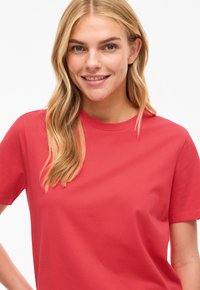 Nuori nainen, jolla on pitkät vaaleat hiukset, hymyilee. Hänellä on päällään yksivärinen punainen lyhythihainen t-paita valkoista taustaa vasten.