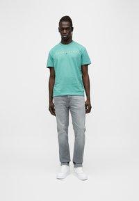 Κοντομάνικο τιρκουάζ μπλουζάκι με το λογότυπο 'TOMMY JEANS' σε κίτρινο, συνδυασμένο με ανοιχτό γκρι τζιν παντελόνι και λευκά αθλητικά παπούτσια, που στέκεται σε απλό φόντο.