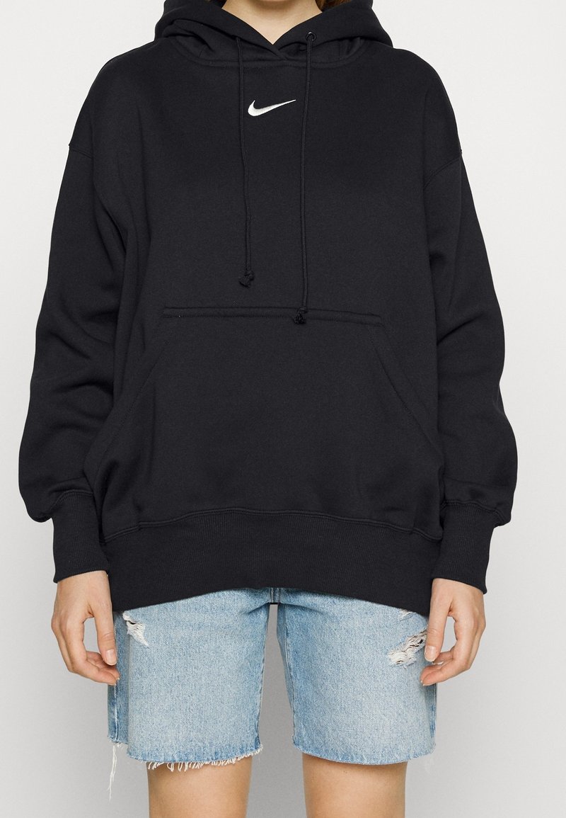 Svart bomullshuvtröja med en framficka, dragsko för huvan och vit Nike-logga. Matchad med slitna denimbyxor.