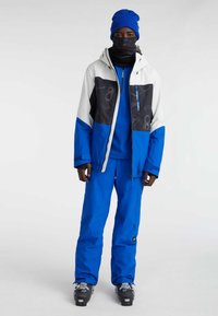 Blauwe skijas met witte en zwarte accenten, gecombineerd met bijpassende blauwe broeken, een blauwe beanie en een zwarte nekwarmer. Inclusief sneeuwlaarzen.