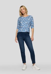 Langärmliges Oberteil mit blauem Muster und Rundhalsausschnitt, kombiniert mit dunklen Skinny Jeans und weißen Sneakers. Glattes Material mit lebhaftem Design.