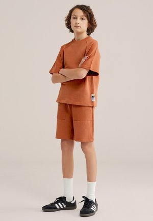 Otrok s kodrastimi lasmi, oblečen v orančasto kratko majico in hlače, bele nogavice ter črne Adidas superge, stoji z rokami prekrižanimi.