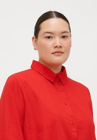 Camicia rossa a bottoni con colletto strutturato e maniche corte, realizzata in tessuto liscio e con una vestibilità rilassata. Design minimalista senza motivi.