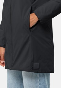 Manteau noir long avec une texture lisse, doté d'une poche latérale et d'un patch logo au bas. Porté sur un denim bleu clair.