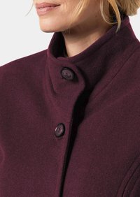 Burgunderfarbener Wollmantel mit hohem Kragen, schwarzen Knöpfen und weicher Textur. Eine Nahaufnahme betont die Knopfdetails und die Stoffzusammensetzung.