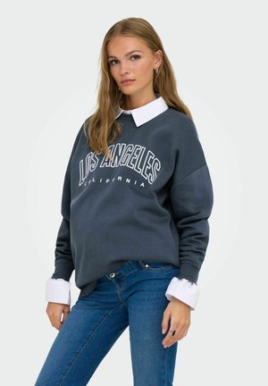 Tumehall sweatshirt, millel on tekst "LOS ANGELES CALIFORNIA", ülepaisutatud lõige, ribakraed, valge kaelusega särke peal. Näha on sinised teksapüksid.