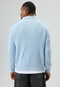 Man met gevlochten haar, gekleed in een lichtblauwe gebreide trui over een wit overhemd en donkere spijkerbroek, met zijn rug naar de camera tegen een effen achtergrond.