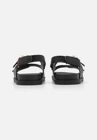 Sandálias pretas com tiras ajustáveis, sola plana e design minimalista; feitas de material suave com um simples detalhe de fivela.