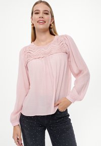 Blusa rosa chiaro con dettagli in pizzo testurizzato sulla parte superiore, maniche lunghe e vestibilità comoda. Abbinata a jeans scuri con pois bianchi.