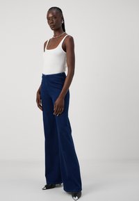 Fehér bordás ujjatlan felső sötétkék bővülő denim farmernadrággal és fekete hegyes orrú magassarkú cipővel, ezüst kiegészítőkkel. A modell egyenesen áll.