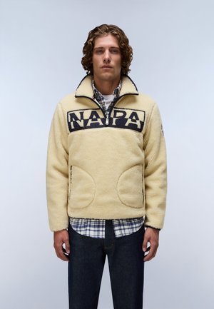Man met krullend haar draagt een beige fleecejack met een halve rits en zwarte tekst over de borst, gedragen over een geruit overhemd en donkere spijkerbroek.