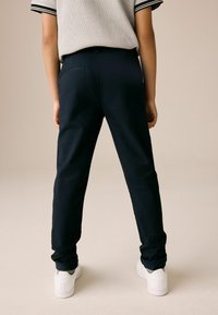 Marineblå bukser med slim fit og glat tekstur, der har en baglomme og er parret med hvide sneakers. Modellen står med ryggen til.