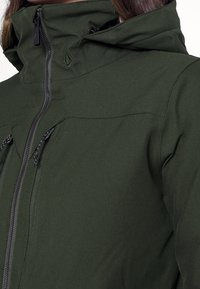 Giacca da esterno verde con colletto alto, chiusura zip e cordini regolabili sul cappuccio. Il materiale è apparentemente testurizzato e durevole.