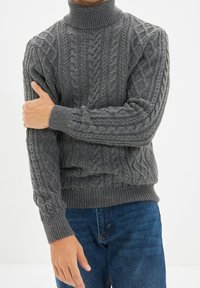 Maglione grigio a coste con collo alto e polsini e orlo a coste, caratterizzato da un motivo testurizzato. Indossato con jeans blu.