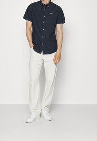 Navy niebieska koszula z krótkim rękawem zapinana na guziki, wykonana z bawełny, z logo na piersi; w połączeniu z kremowymi spodniami z lnu i białymi sneakerami.