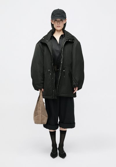 Black padded jacket, black shirt, rolled black pants, dark socks, and pointed shoes. Holds a tan suede bag; features a peaked cap.