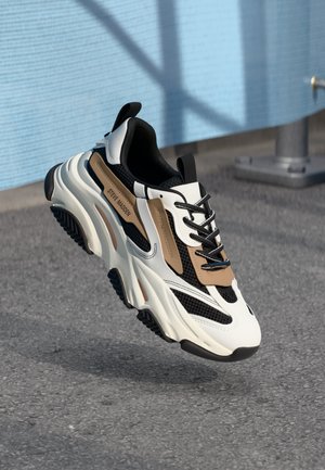 Sneaker bianchi e neri con accenti marroni, materiali in rete e sintetici, punta rotonda, suola testurizzata e lacci neri.