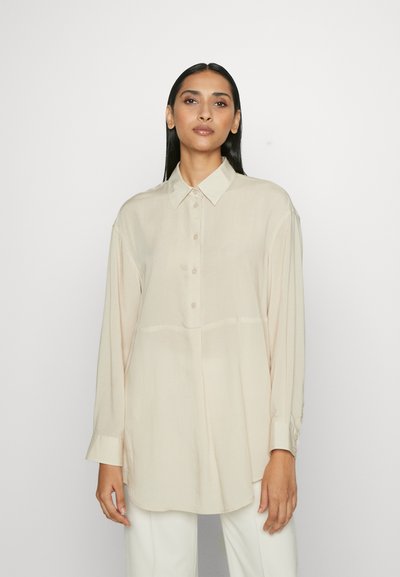 Camicia a maniche lunghe color beige chiaro con chiusura a bottoni, dettagli con pieghe e tessuto morbido e liscio. Presenta un colletto classico e una vestibilità comoda.