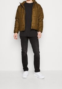 Chaqueta acolchada de color verde oliva con cierre de cremallera y capucha, que presenta un diseño acolchado. Combinada con una camisa negra y vaqueros oscuros, completada con zapatillas blancas.