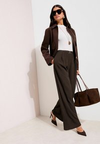 Bruine op maat gemaakte jas over een wit topje, gecombineerd met wijde, geribbelde broekspijpen. Accessoires zijn onder andere een zonnebril, een lange ketting en een bruine tas.