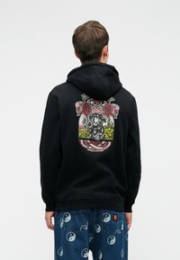 Mustahuppari, jossa on graafinen suunnittelu, joka esittää ruusuja ja hahmoa kukkivassa ympäristössä selässä. Puuvillamateriaali, rento istuvuus.