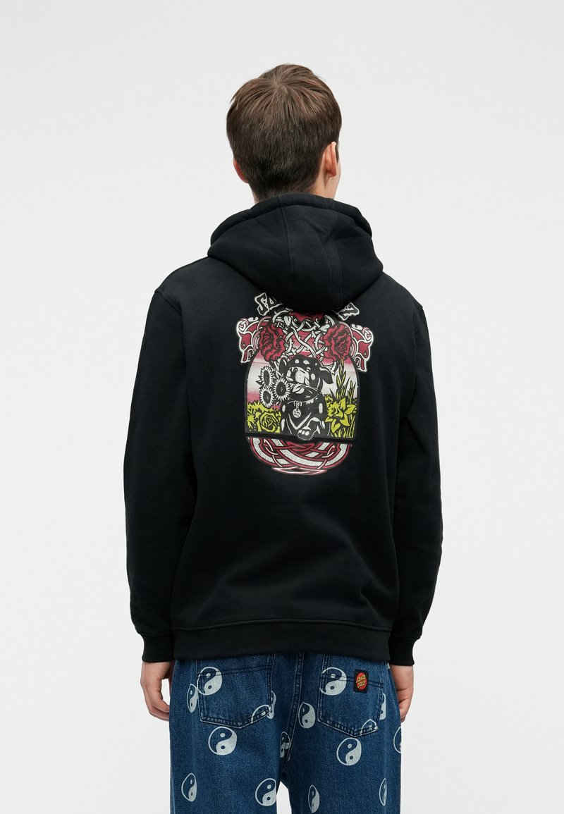 Mustahuppari, jossa on graafinen suunnittelu, joka esittää ruusuja ja hahmoa kukkivassa ympäristössä selässä. Puuvillamateriaali, rento istuvuus.