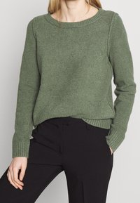 Sweter w kolorze oliwkowym z dzianiny, z okrągłym dekoltem, ściągaczami przy mankietach i u dołu. Zaprezentowany w połączeniu z czarnymi spodniami. Miękka struktura i luźny krój.