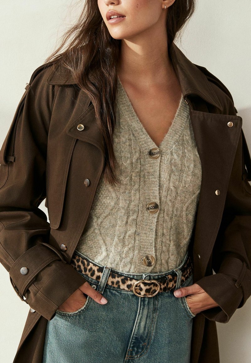 Bruine trenchcoat met knoopdetails over een lichtgrijze kabelgebreide trui. Heeft een riem met luipaardprint en denim met een vervaagde afwerking.