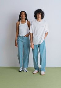 Deux mannequins se tiennent côte à côte. La femme porte un débardeur blanc ajusté et un jean clair taille haute. L'homme porte un t-shirt blanc surdimensionné et un jean clair ample.
