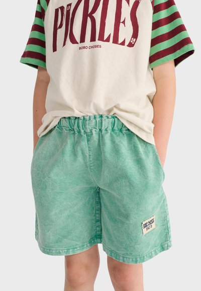 Enfant portant un short élastique taille haute vert délavé à l'acide et un T-shirt blanc avec des manches rayées vertes et bordeaux et le texte "PICKLES".
