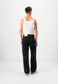 Top blanco sin mangas, jeans negros desgastados con acentos de strass y un parche amarillo horizontal en la cinturilla trasera. Calzado casual visible.