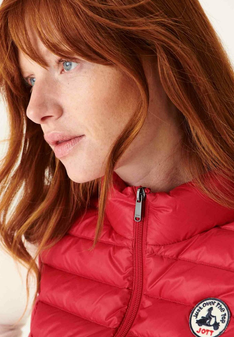 Red, padded vest with a high collar, featuring a zip closure and circular logo patch with text. Smooth fabric and quilted design.