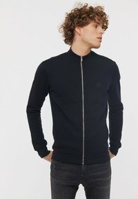 Veste zippée noire avec un col haut, des poignets côtelés et une coupe ajustée. Présente un petit logo sur la poitrine et est fabriquée en tissu lisse et extensible.
